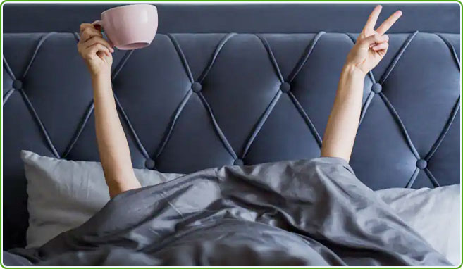 Groupon Coupons Hotwire a person in bed head under the covers with just their arms showing. One hand is flashing a peace sign, the other has a coffee mug